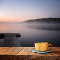 Front image of coffee cup over wooden table in front of calm foggy lake view at sunset