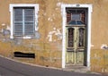 Front of an old abandoned yellow house with blue shuttered windows and locked green wooden door with flaking peeling paint on