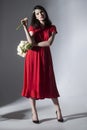 Front photo of an attractive young brunette woman in red dress holding a bouquet of daffodils, isolated grey background.