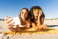 Front portrait of two cute girls with sunglasses in summer vacation using smartphone taking a selfie lying on sea sand of a