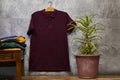 Front side of male garnet cotton t-shirt hanging on gray grunge marble wall with cactus pot and stacked shirts. Royalty Free Stock Photo