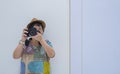 Front view of Asian female tourist photographing with digital camera while leaning against the wall inside of white room Royalty Free Stock Photo