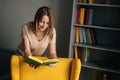 Front view of attractive blonde woman standing near yellow chair reading paper book in dark room with modern interior, Royalty Free Stock Photo