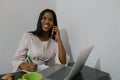Front view of an attractive young African woman working on a laptop and smiling while talking on her cell phone Royalty Free Stock Photo