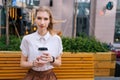 Front view of attractive young woman in casual clothes sitting on bench holding paper cup of takeaway coffee looking at Royalty Free Stock Photo