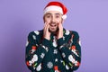 Front view of bearded guy dresses santa hat and fanny sweaterv with snowman looking astonished, keeping hands near his mouth,