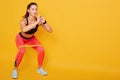 Front view of beautiful slim muscular female with resistance band performing squats isolated over yellow background, sporty woman