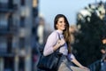 Front view of beautiful trendy young woman smiling and wearing casual wear sitting on a metallic fence in the street while looking