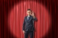 Front view of businessman standing in spotlight against red stage curtain looking at camera and holding out hand with