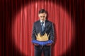 Front view of businessman standing in spotlight against red stage curtain looking down at gold crown on blue pillow