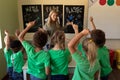 Female school teacher pointing to a recycling poster with a group of schoolchildren raising their ha