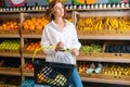 Front view of cheerful young woman customer holding basket of fruits standing at fruit and vegetables section of grocery Royalty Free Stock Photo