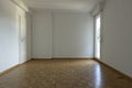 Front view of an empty room with parquet flooring and white walls. On the left is a window letting in a lot of natural light, on Royalty Free Stock Photo