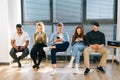 Front view of group five young diverse ethnicity candidates using mobile phones while waiting job interview in modern