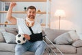 Front view. Holding soccer ball and watching sport match. Man with crutches is at home indoors. Having injuries Royalty Free Stock Photo