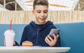 Teenager boy sits at table in cafe, drinks milkshake, eats donut, holds smartphone in his hand. Boy plays mobile games. Royalty Free Stock Photo