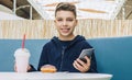 Teenager boy sits at table in cafe, drinks milkshake, eats donut, holds smartphone in his hand. Boy plays mobile games. Royalty Free Stock Photo