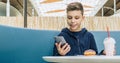 Teenager boy sits at table in cafe, drinks milkshake, eats donut, holds smartphone in his hand. Boy plays mobile games. Royalty Free Stock Photo