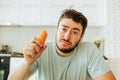 Front view looking at the camera a young man is sad holding a raw carrot in his hand.