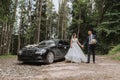 Front view of a married bride and groom wearing festive clothes standing against a black car Royalty Free Stock Photo