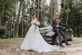 Front view of a married bride and groom wearing festive clothes standing against a black car Royalty Free Stock Photo