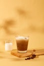 Front view of a milk coffee cup is placed on a wooden platform, next to it is milk in a beaker and coffee beans on a shaded beige