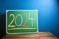 Front view of the number 2024 written in chalk on green chalkboard placed on wooden table against flat blue wall. New year and