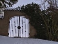 View of old stone wall with iron gate and decorated lattice on winter day with snow-covered path, climbing plant and bare bush. Royalty Free Stock Photo