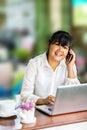 Front view portrait of Asian businesswoman sitting at long table in coffee shop and using laptop computer and talking on cell phon Royalty Free Stock Photo