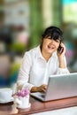 Front view portrait of Asian businesswoman sitting at long table in coffee shop and using laptop computer and talking on cell phon Royalty Free Stock Photo