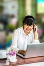 Front view portrait of Asian businesswoman sitting at long table in coffee shop and using laptop computer and talking on cell phon Royalty Free Stock Photo