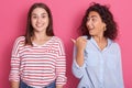 Front view portrait of charming astonish woman standing and pointing with her thumb to her smiling female wearing striped casual