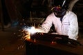 Front view of professional welder in white uniform with protective helmet welding steel with spark in workshop. Industrial worker