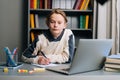 Front view of pupil boy making notes in copybook with pen sitting at desk with laptop computer looking at camera. Royalty Free Stock Photo