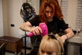 Front view of a red-haired woman professional hairdresser with tattooed arms in a strict black uniform combing, straightening and Royalty Free Stock Photo