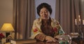 Front view of senior Caucasian woman in shawl sitting at the table with candles and shuffling cards. Positive old witch