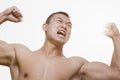 Front view of shirtless, angry, roaring young man flexing his muscles with arms raised and looking away