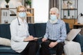 Front view shot of young woman nurse doctor in mask, consulting senior gray haired woman during homecare medical visit Royalty Free Stock Photo