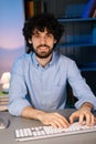 Front view of smiling bearded young man looking at camera and typing online message using computer. Royalty Free Stock Photo