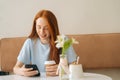 Front view of smiling young woman using smartphone chatting with friends and drinking hot coffee sitting at desk in cozy Royalty Free Stock Photo