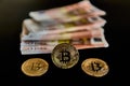 Front view of three Bitcoin coins, next to a group of Euro banknotes, on a black background. Concept of money, power, economics,