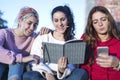 Front view of three girls sitting on ground outdoors. One of them using mobile phone while the rest looking at a Book Royalty Free Stock Photo