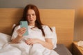 Front view of tired ill red-haired young woman having online video call via smartphone lying in bed at home. Sick Royalty Free Stock Photo