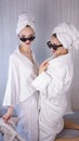 Front view of two smiling female friends with towels and in bathrobes sitting on bed and doing pedicure. Cute happy young girls Royalty Free Stock Photo