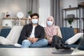 Front view of young arabic muslim couple in medical masks sitting on soft sofa at home. Isolation, desease, infection