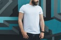 Front view. Young bearded hipster man dressed in white t-shirt is stands against wall with graffiti. Mock up. Royalty Free Stock Photo