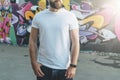 Front view. Young bearded hipster man dressed in white t-shirt is stands against wall with graffiti. Mock up.