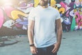 Front view. Young bearded hipster man dressed in white t-shirt is stands against wall with graffiti. Mock up.