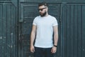 Front view. Young bearded hipster man dressed in white t-shirt and sunglasses is stands against dark wood wall.