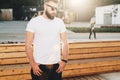 Front view. Young bearded hipster man dressed in white t-shirt and sunglasses is stands on city street. Mock up. Royalty Free Stock Photo
